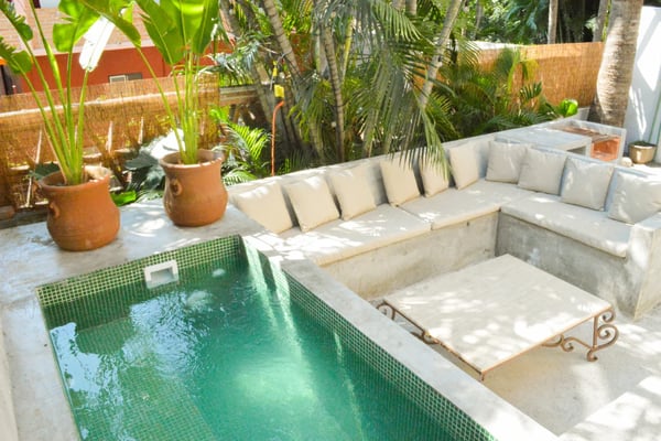 Sunlit courtyard with green-tiled pool, built-in concrete seating with cream cushions, terracotta planters, and tropical palm trees