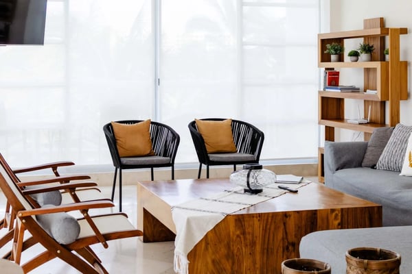 Bright living room with wooden lounge chairs, black rope seating, marble coffee table, gray sofa, and wooden shelving unit with potted plants